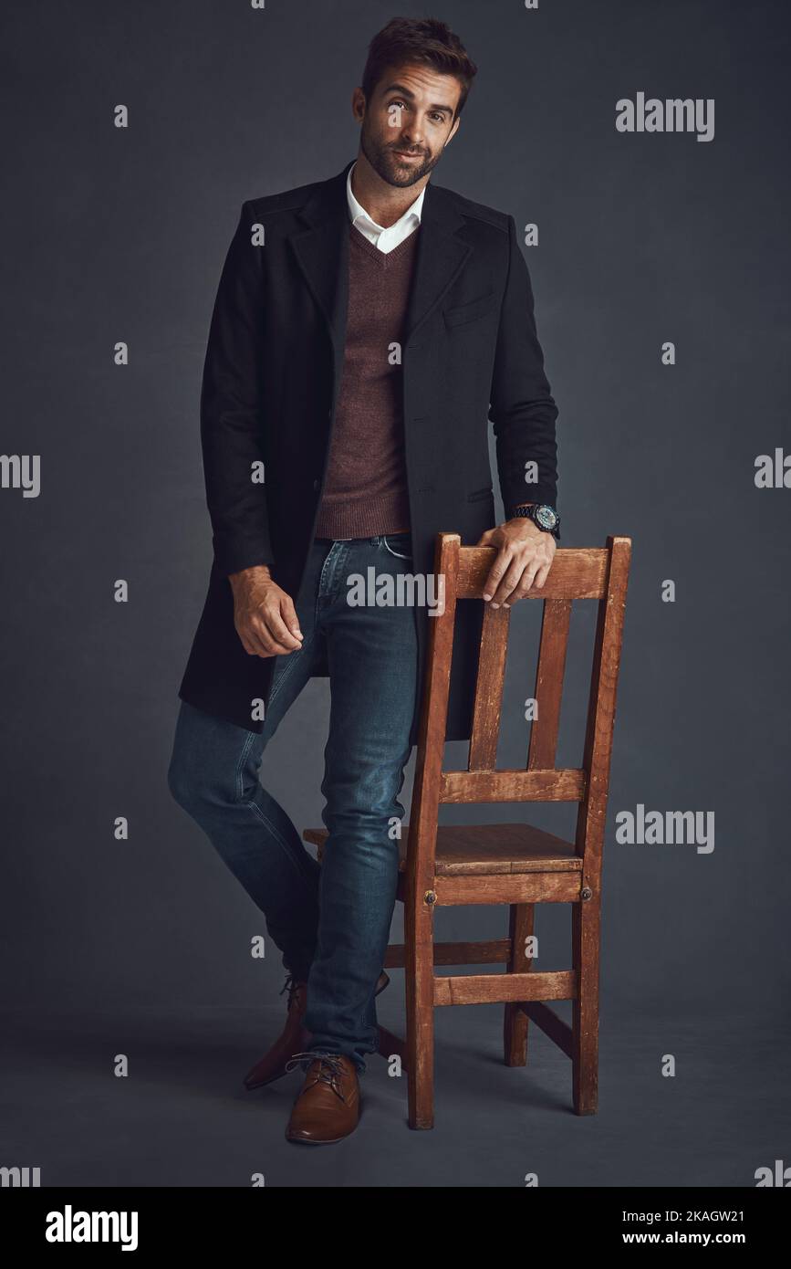 Lo stile è facile per lui. Studio ritratto di un giovane uomo elegantemente vestito in piedi accanto a una sedia su uno sfondo grigio. Foto Stock