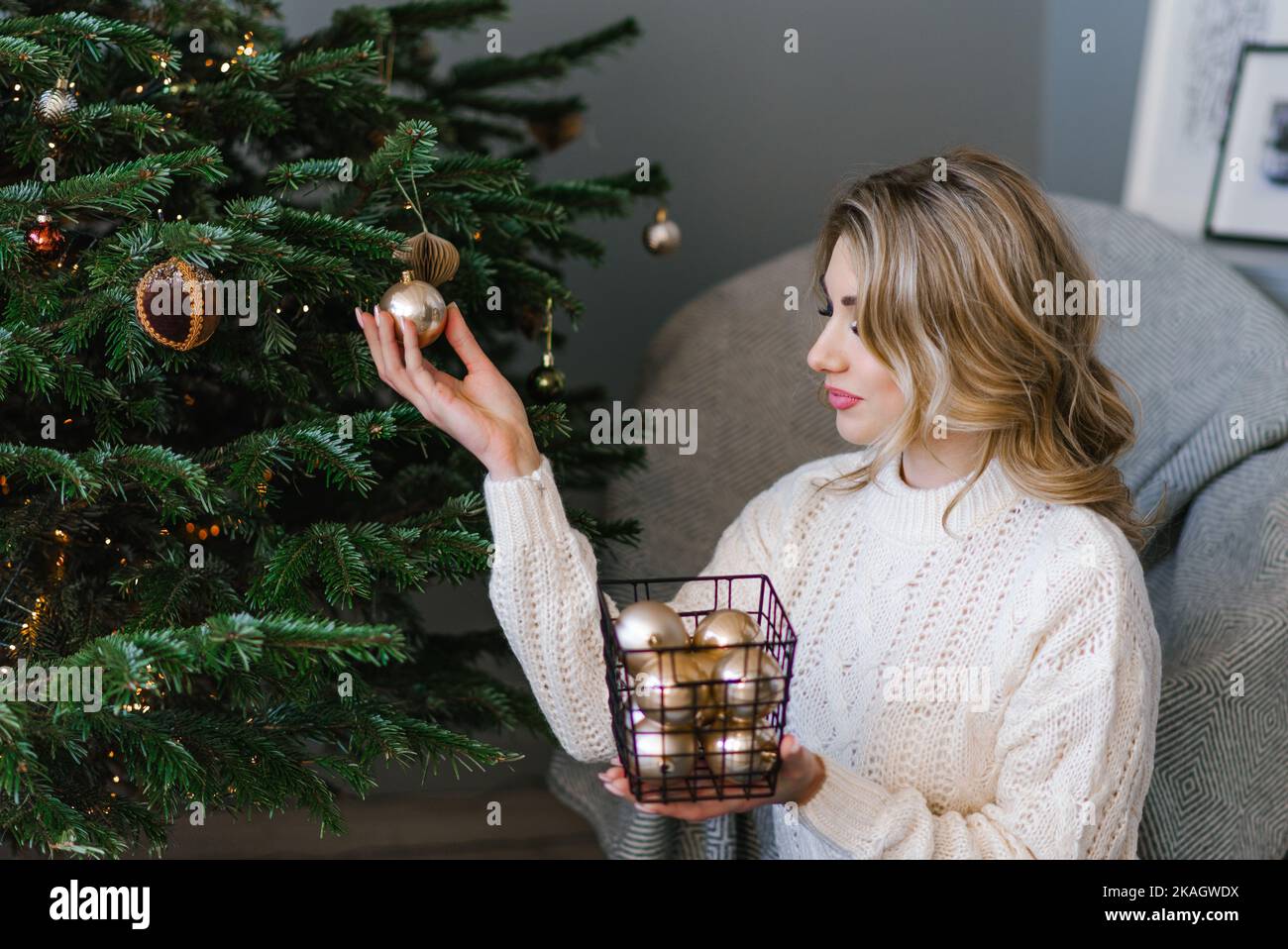 Bella giovane donna in un maglione accogliente e jeans decorare un albero di Natale con palloncini da un cesto a casa Foto Stock