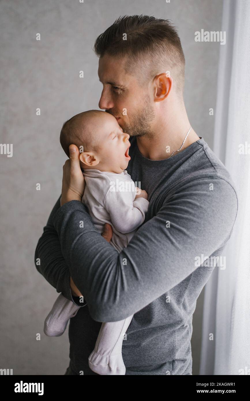 Un giovane padre tiene fra le braccia il figlio piccolo e lo bacia, il bambino urla Foto Stock