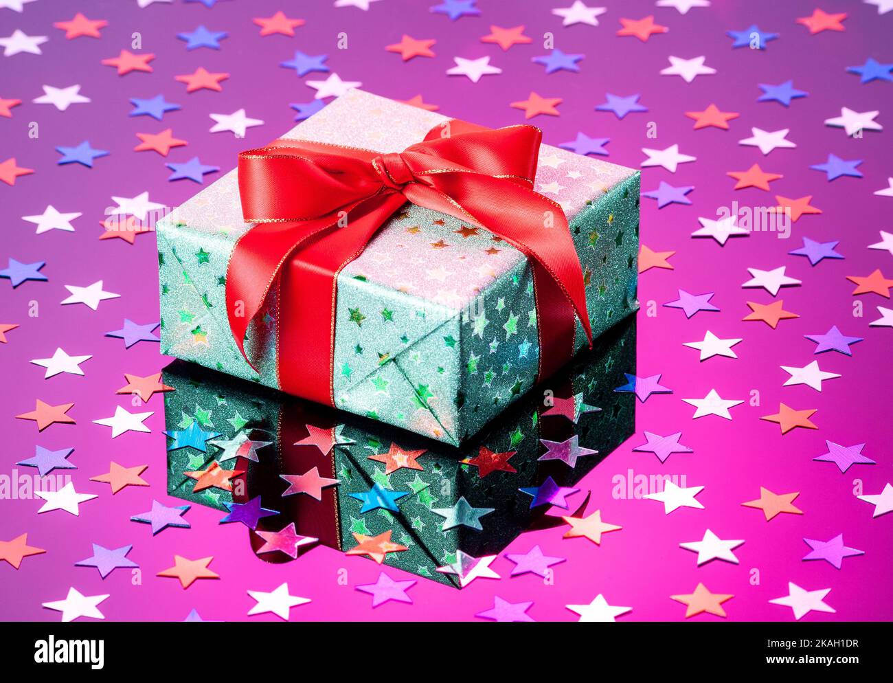 Confezione regalo verde singola con fiocco a nastro rosso su superficie riflettente viola con stelle glitterate. Confezione regalo avvolta in carta lucida verde con chiusura nastro rosso Foto Stock