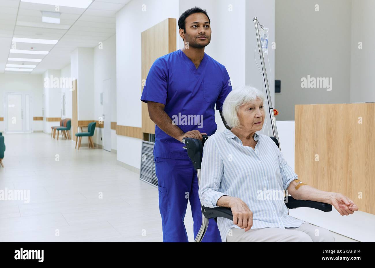 Infermiera maschile indiana che spinge la sedia a rotelle con la donna anziana in corridoio ospedale. Assistenza agli anziani Foto Stock