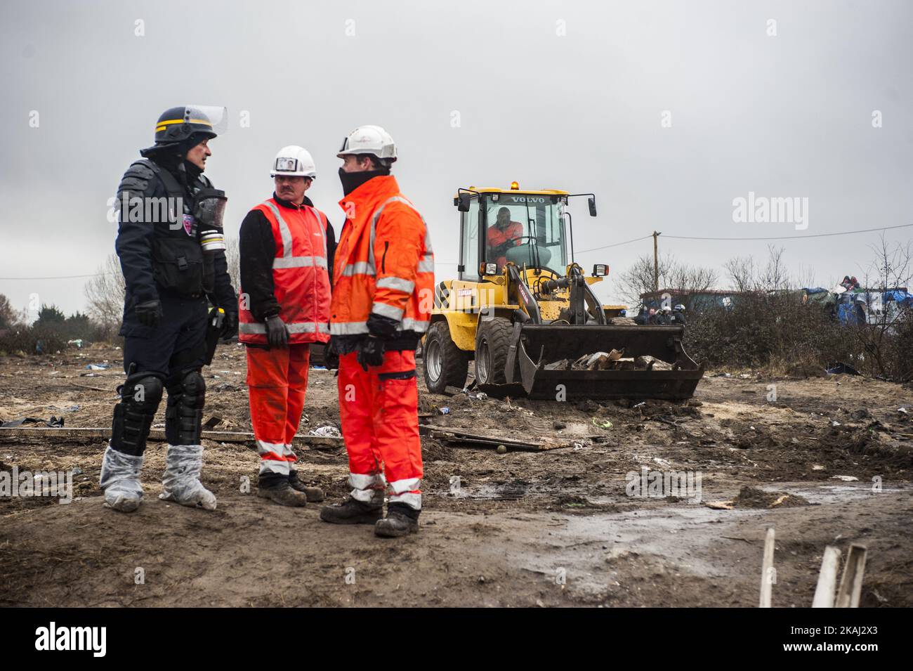 Un lavoratore discute con la polizia durante la distruzione della parte meridionale della giungla. Un retroescavatore raccoglie capanne smantellate. A Calais, Francia, il 1 marzo 2016. Secondo quanto riferito, i funzionari vanno porta a porta nel campo dei migranti della giungla a Calais per cercare di convincere le persone a lasciare il sito e a essere delocalizzate come richiedenti asilo in Francia. Si dice che anche le ONG e gli attivisti siano a disposizione per fornire una controargomentazione giuridica. Le squadre francesi di demolizione hanno iniziato a smantellare le capanne all'inizio di questa settimana, ma questo è stato incontrato con resistenza, con la polizia tumultuata che è stata costretta a sparare gas lacrimogeni contro gli immigrati che gettano lo sto Foto Stock