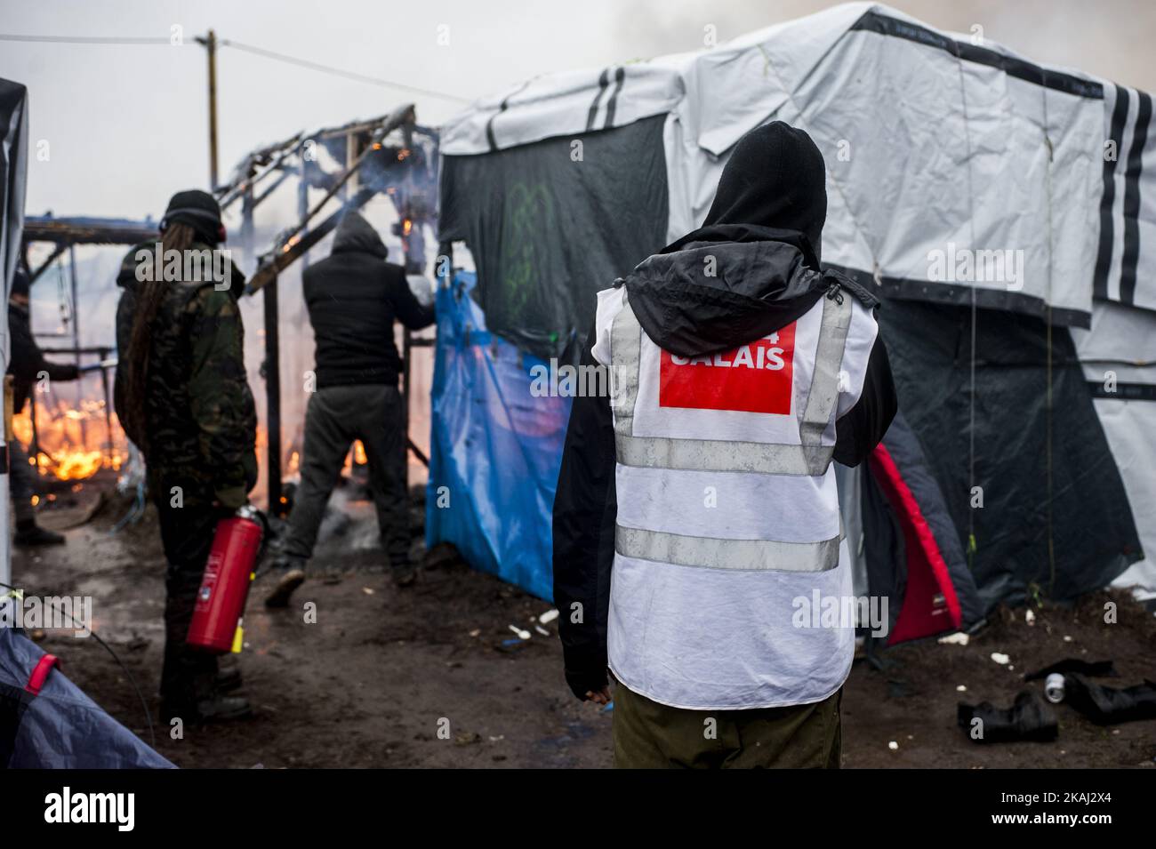 Un attivista DI CURA 4 CALAIS aiuta i migranti a spegnere l'incendio durante l'evacuazione della zona a sud della giungla. A Calais, in Francia, il 1 marzo 2016. Secondo quanto riferito, i funzionari vanno porta a porta nel campo dei migranti della giungla a Calais per cercare di convincere le persone a lasciare il sito e a essere delocalizzate come richiedenti asilo in Francia. Si dice che anche le ONG e gli attivisti siano a disposizione per fornire una controargomentazione giuridica. Le squadre francesi di demolizione hanno iniziato a smantellare capanne all'inizio di questa settimana, ma questo è stato incontrato con resistenza, con la polizia antisommossa che è stata costretta a sparare gas lacrimogeni a causa dei migranti che gettano s. Foto Stock