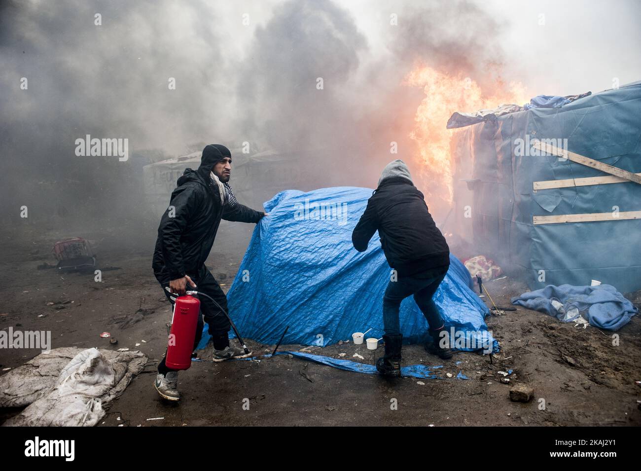 Un migrante e benevole cercando di rimuovere una tenda vicino al fuoco in modo che non si diffonda al resto della cabina durante l'evacuazione della zona sud della giungla. A Calais, Francia, il 1 marzo 2016. Secondo quanto riferito, i funzionari vanno porta a porta nel campo dei migranti della giungla a Calais per cercare di convincere le persone a lasciare il sito e a essere delocalizzate come richiedenti asilo in Francia. Si dice che anche le ONG e gli attivisti siano a disposizione per fornire una controargomentazione giuridica. Le squadre francesi di demolizione hanno iniziato a smantellare capanne all'inizio di questa settimana, ma questo è stato incontrato con resistenza, con la polizia tumulto che è stata costretta a f Foto Stock