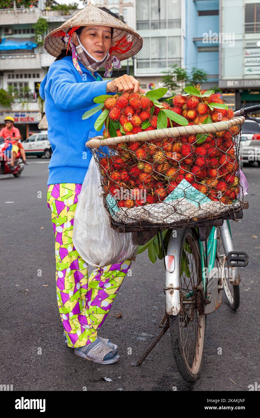 Signora vietnamita che indossa cappello di bambù, vendendo il rambutan da una bicicletta nel centro di ho Chi Minh City, Vietnam Foto Stock