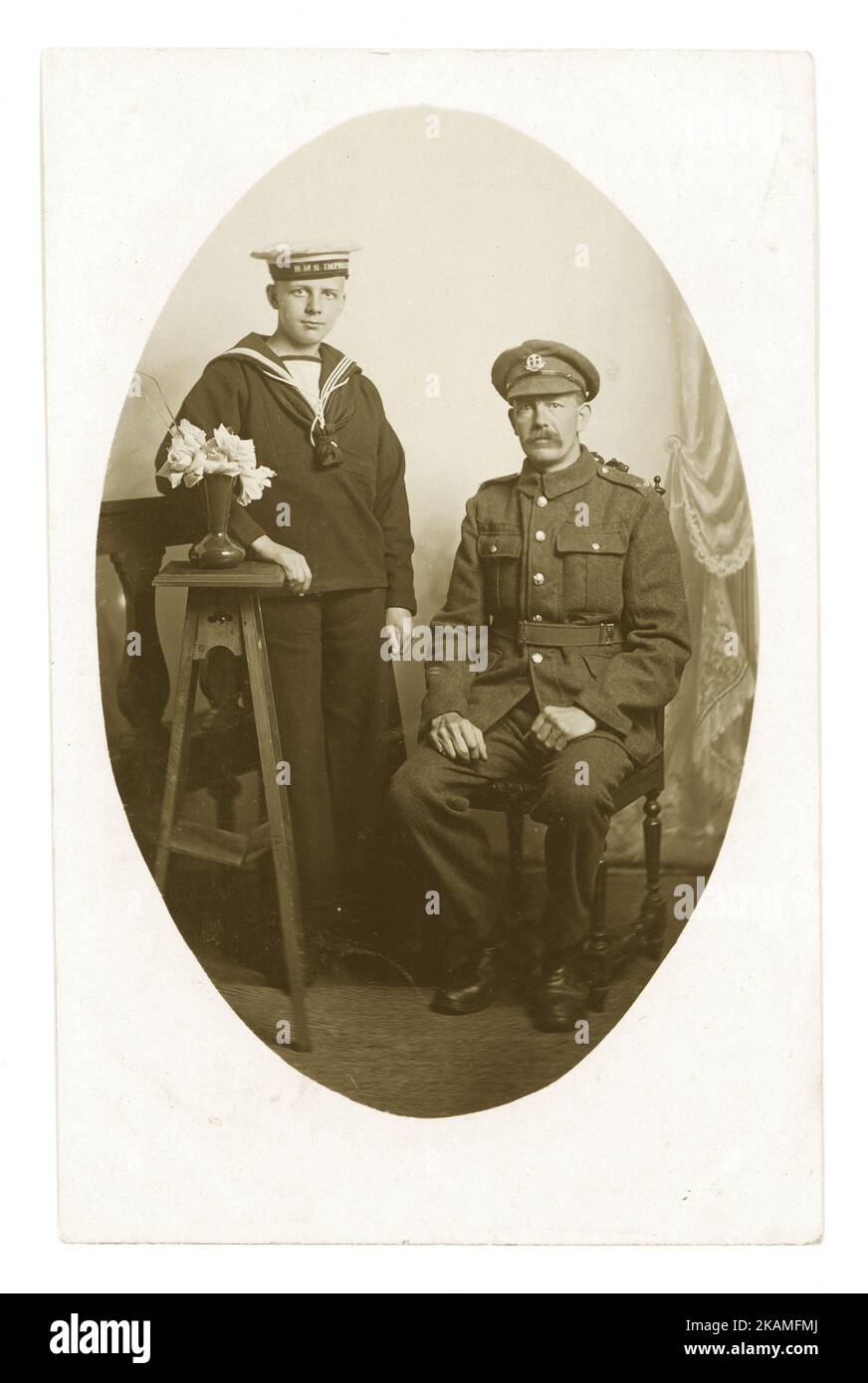 WW1 era cartolina studio ritratto di padre e figlio guardando solenne. Il padre è con il reggimento Middlesex (badge su cappuccio) e il figlio è un cadetto navale su HMS imprendibile, una nave da addestramento (HMS imprendibile su cappuccio), Middlesex, S.E. Inghilterra vicino a Londra, circa 1916 Foto Stock