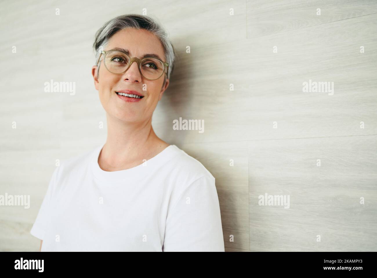 Donna d'affari allegra guardando via pensieroso mentre si appoggia contro una parete in un ufficio. Imprenditrice femminile matura in piedi da sola in un lavoro moderno Foto Stock