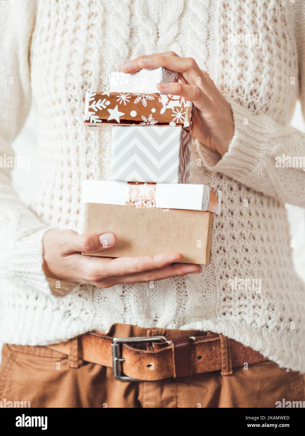 Donna in maglia bianca con motivo scandinavo e pantaloni chino marroni con cintura in pelle. Pila di regali di Natale in mano. Foto Stock