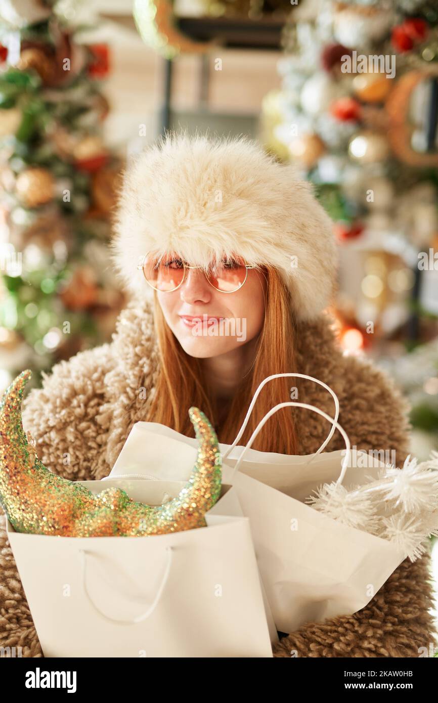 Attraente ragazza caucasica in caldo abbigliamento outerwear e occhiali da sole con borse per la spesa piena di decorazione di Natale in negozio o centro commerciale. Shopping di Capodanno o tema di consumismo Foto Stock