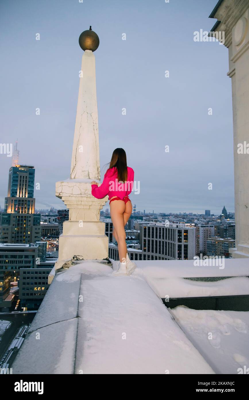Ragazza che indossa maglione rosa, tanga e sneakers si trova vicino al pilastro sul tetto di un grattacielo che guarda fuori la città, in cima al concetto di mondo Foto Stock