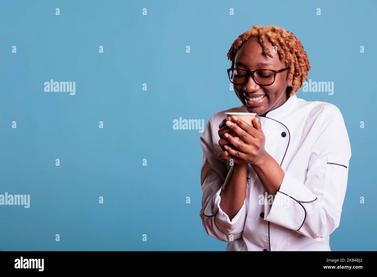 Rilassato cuoca femminile gustando una tazza di caffè preferito durante l'ora di pranzo. Chef professionista afroamericano che indossa uniforme da cucina su sfondo blu in studio shot. Foto Stock