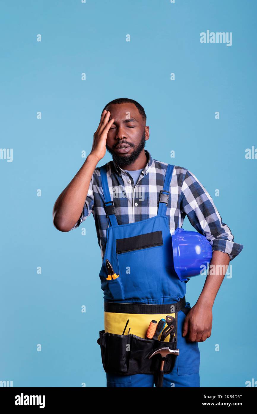 Costruttore stressato in uniforme e stanco elmetto strofinando la sua testa sensazione di affaticamento e mal di testa. Impiegato di costruzione frustrato in dolore severo dovuto lavoro duro e mancanza di riposo contro lo sfondo blu. Foto Stock