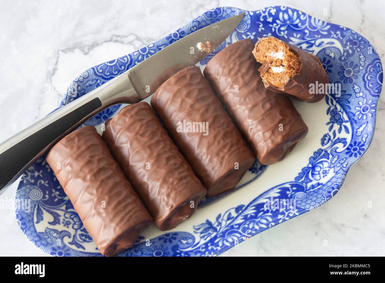 Rotoli di spugna al cioccolato individuali su un piatto blu e bianco a motivi geometrici con un coltello da portata. Su uno sfondo di marmo bianco Foto Stock