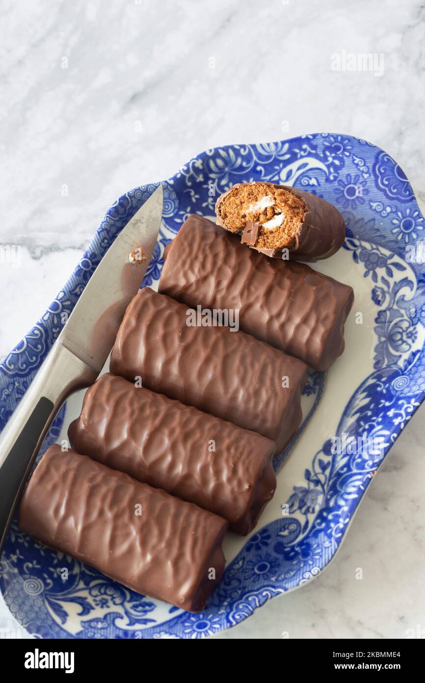 Rotoli di spugna al cioccolato individuali su un piatto blu e bianco a motivi geometrici con un coltello da portata. Su uno sfondo di marmo bianco Foto Stock