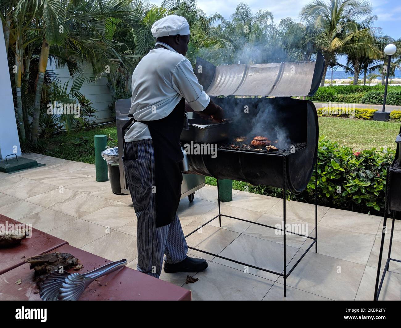 Uno chef cuoce il pollo cretino su un tradizionale barbecue a tamburo di olio davanti alle palme, un prato e il mare. Cultura giamaicana. Foto Stock