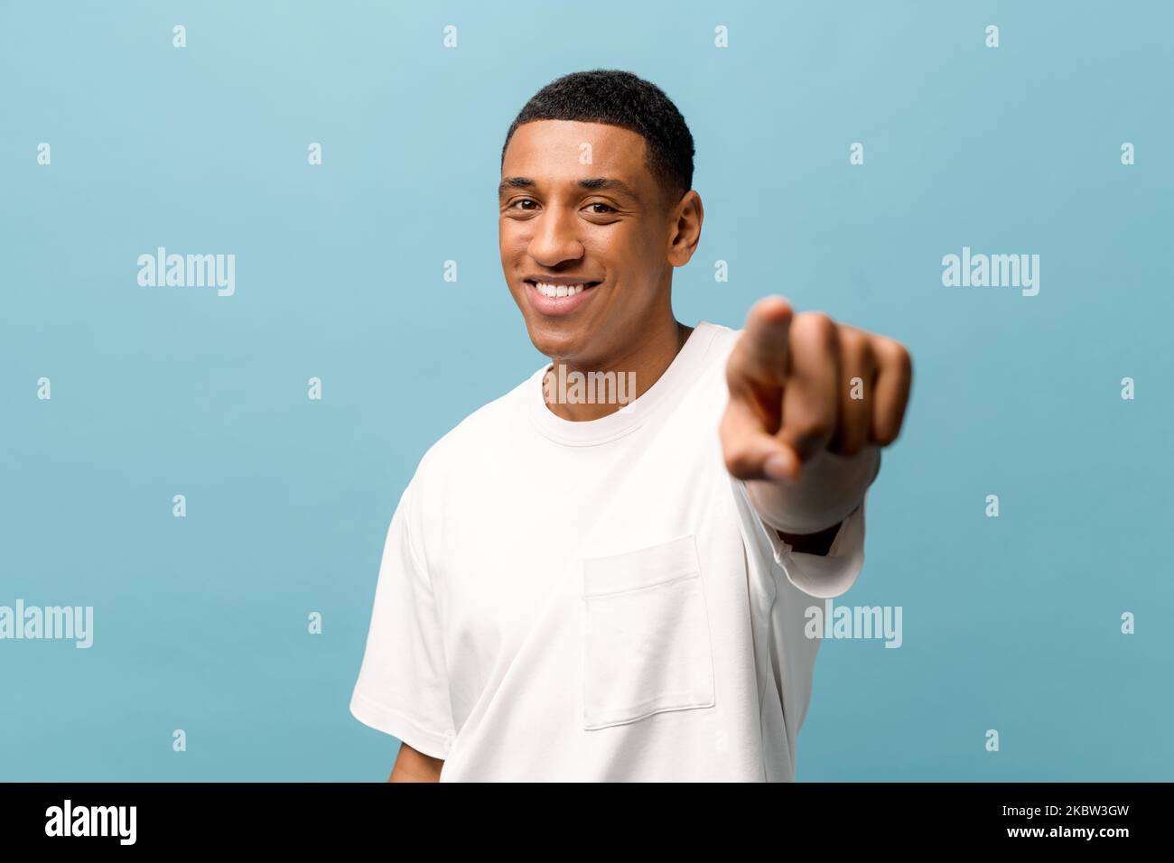 Attraente giovane ragazzo afro-americano allegro indossare bianco casual t-shirt punti indice dito alla fotocamera, scegliendo si isolato su sfondo blu, felice uomo millenario mostrando direzione alla fotocamera Foto Stock