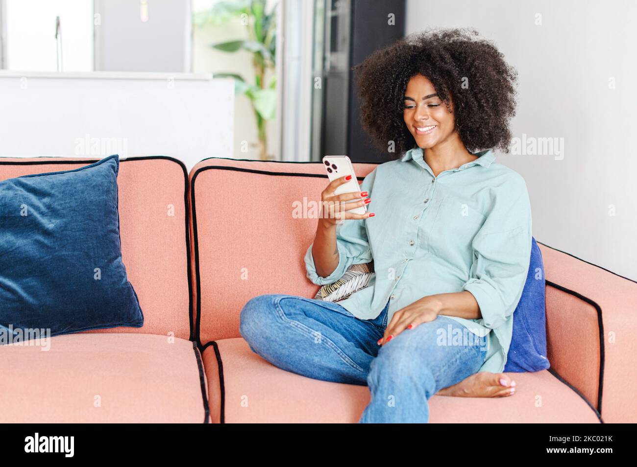 Ritratto di una donna sorridente seduta sul divano di casa, utilizzando il telefono cellulare per controllare le reti sociali, digitando un messaggio per la famiglia, visualizzare foto, guardare video Foto Stock