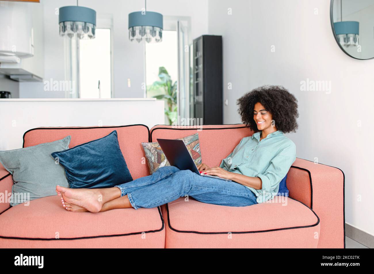 Vista a tutta lunghezza della donna d'affari nera o della freelancer femminile in comodi abiti casual seduti sul divano e lavorando al progetto, digitando e-mail sul computer portatile, scrivendo un articolo Foto Stock