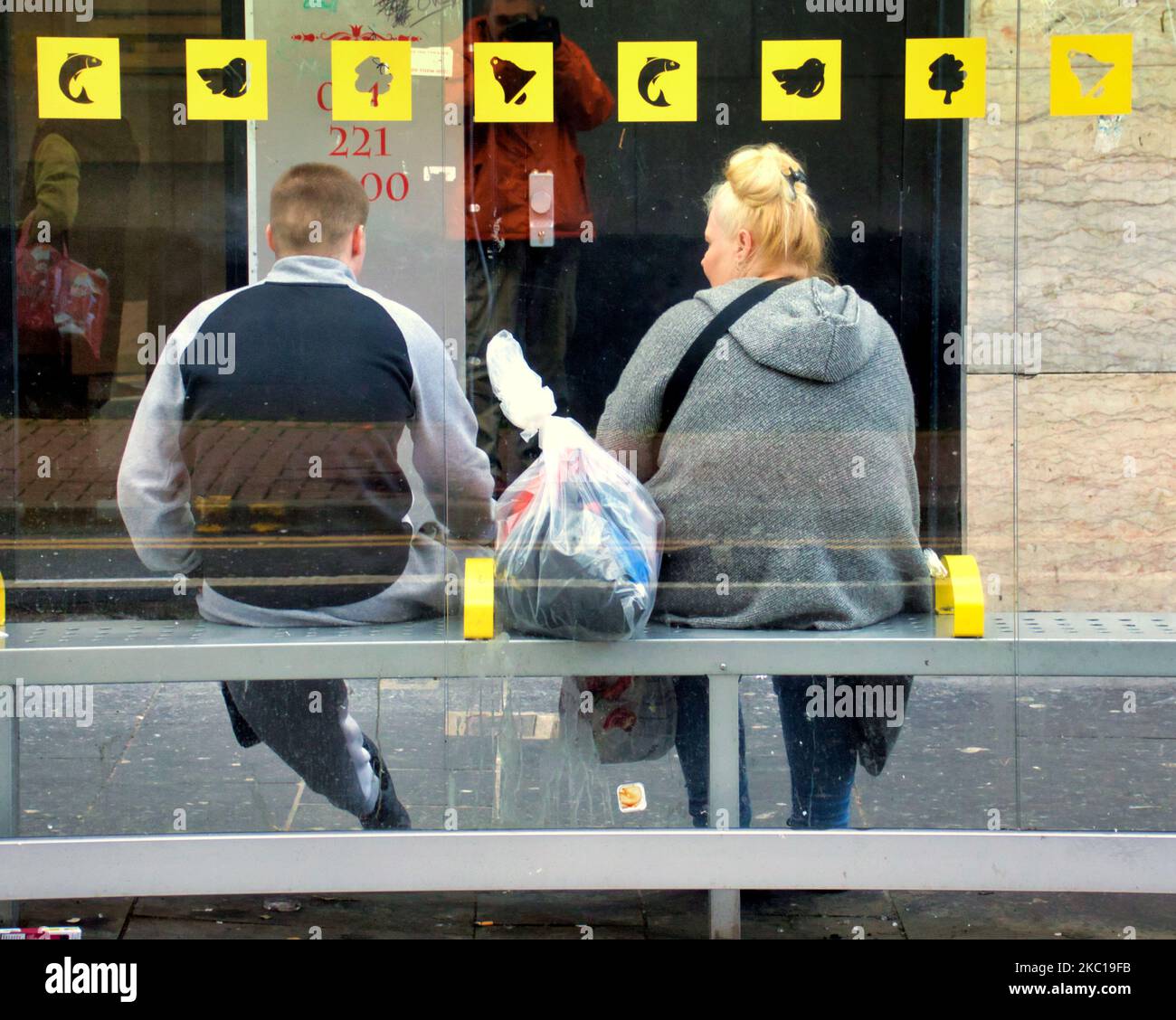 Passeggeri in sovrappeso a una fermata dell'autobus sotto i simboli iconici della città di Glasgow del suo mito Foto Stock