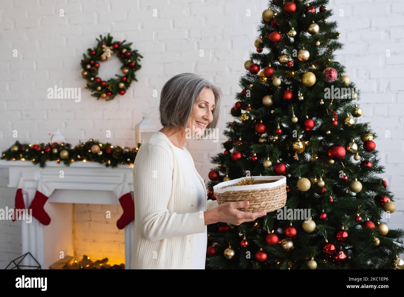 donna allegra e matura che tiene un cesto di vimini vicino all'albero di natale decorato a casa Foto Stock donna allegra e matura che tiene un cesto di vimini vicino all'albero di natale decorato a casa Foto Stock