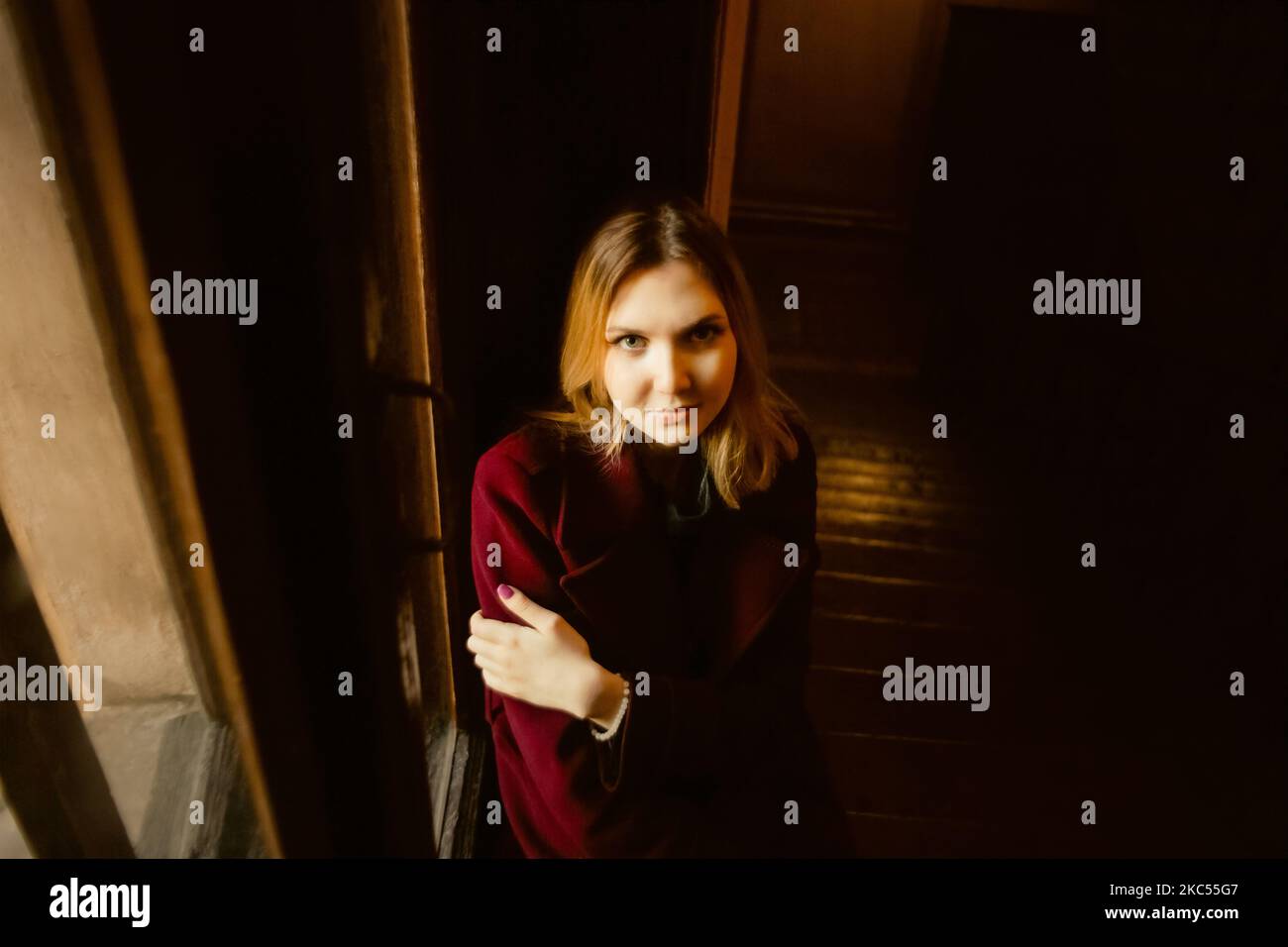 Ritratto di un bellissimo studente biondo in un cappotto di borgogna, che è seduto su un vecchio davanzale vintage vicino alla finestra solo la sera. Foto Stock