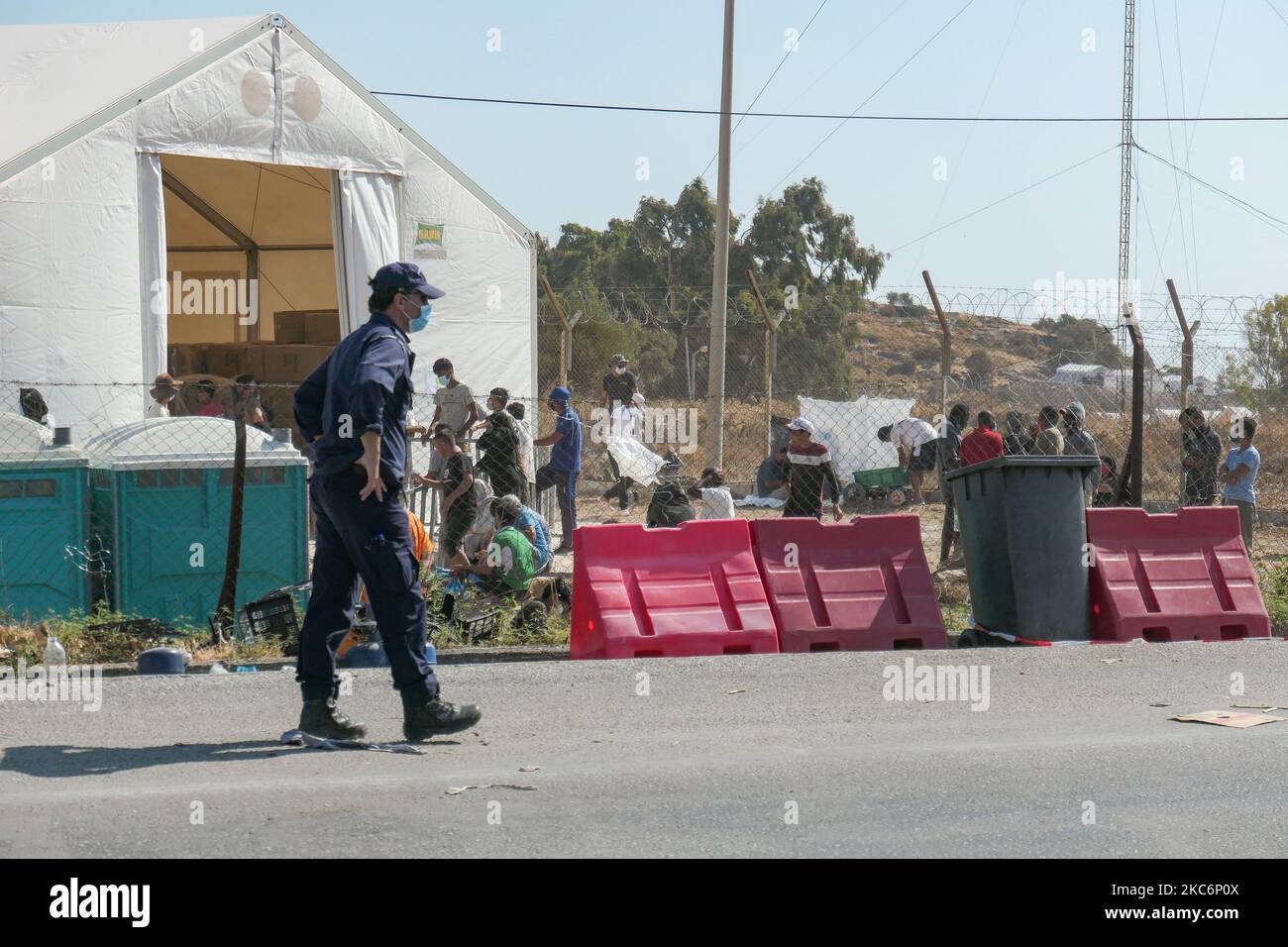 Un ufficiale di polizia che pattugliava al di fuori del nuovo campo temporaneo per rifugiati a Kara Tepe - Mavrovouni con grandi tende bianche con il logo UNHCR o UNICEF e richiedenti asilo in attesa di registrazione e forniture come cibo, letti, vestiti, ecc come visto dall'ingresso principale custodito. Il nuovo campo recintato è un'ex area militare, campo di tiro dell'esercito ellenico, vicino alla città di Mytilene nell'isola di Lesbos Grecia. Il nuovo campo fu creato dopo l'incendio del 9 settembre 2020, quando fu uno dei più grandi campi profughi d'Europa, il campo profughi di Moria (centro di accoglienza, identificazione e registrazione) Foto Stock