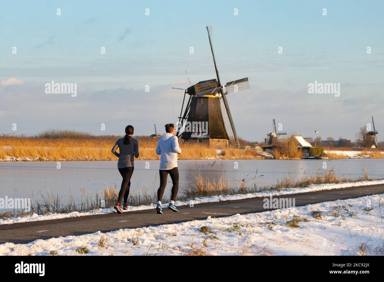 Una coppia come visto in esecuzione a Kinderdijk. Una magica scriglia intorno al tramonto e crepuscolo con il cielo limpido d'inverno ai canali ghiacciati e ai mulini a vento di Kinderdijk nei Paesi Bassi. Il paese sta affrontando il clima artico, le temperature basse severe, le nevicate ed i venti alti dovuto la tempesta Darcy che ha causato i problemi nei trasporti e nella vita di tutti i giorni. Molti dei canali sono stati ghiacciati perché la temperatura è inferiore a zero a causa del clima freddo che a volte raggiunge i -16C, mentre il terreno è coperto di neve. Kinderdijk e i mulini a vento nel polder Alblasserwaard, è una famosa attrazione, un punto di riferimento olandese e. Foto Stock