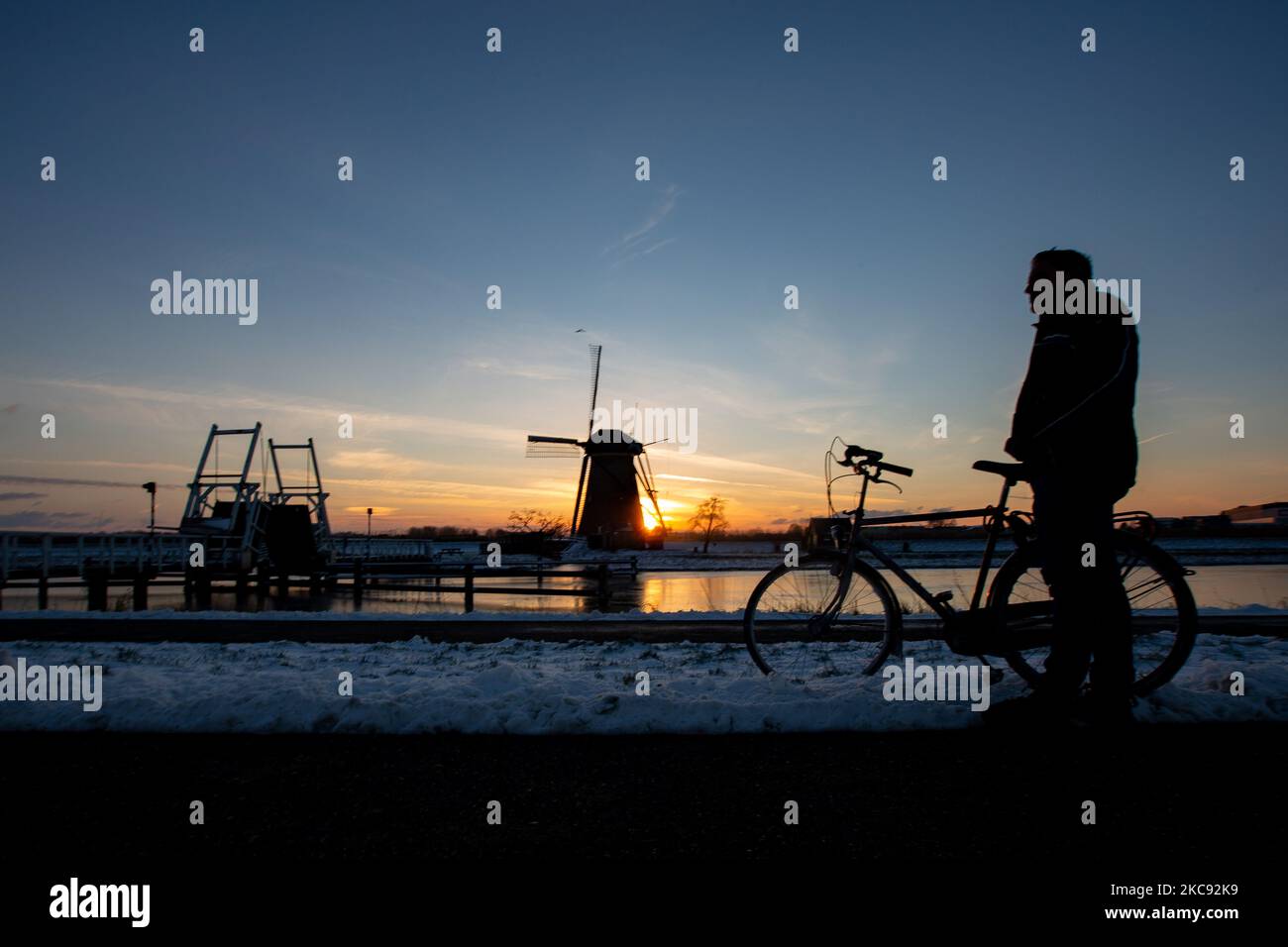 Un uomo si sta godendo il tramonto accanto alla sua bicicletta. Una magica scriglia intorno al tramonto e crepuscolo con il cielo limpido d'inverno ai canali ghiacciati e ai mulini a vento di Kinderdijk nei Paesi Bassi. Il paese sta affrontando il clima artico, le temperature basse severe, le nevicate ed i venti alti dovuto la tempesta Darcy che ha causato i problemi nei trasporti e nella vita di tutti i giorni. Molti dei canali sono stati ghiacciati perché la temperatura è inferiore a zero a causa del clima freddo che a volte raggiunge i -16C, mentre il terreno è coperto di neve. Kinderdijk e i mulini a vento nel polder Alblasserwaard, è una famosa attrazione, una olandese la Foto Stock