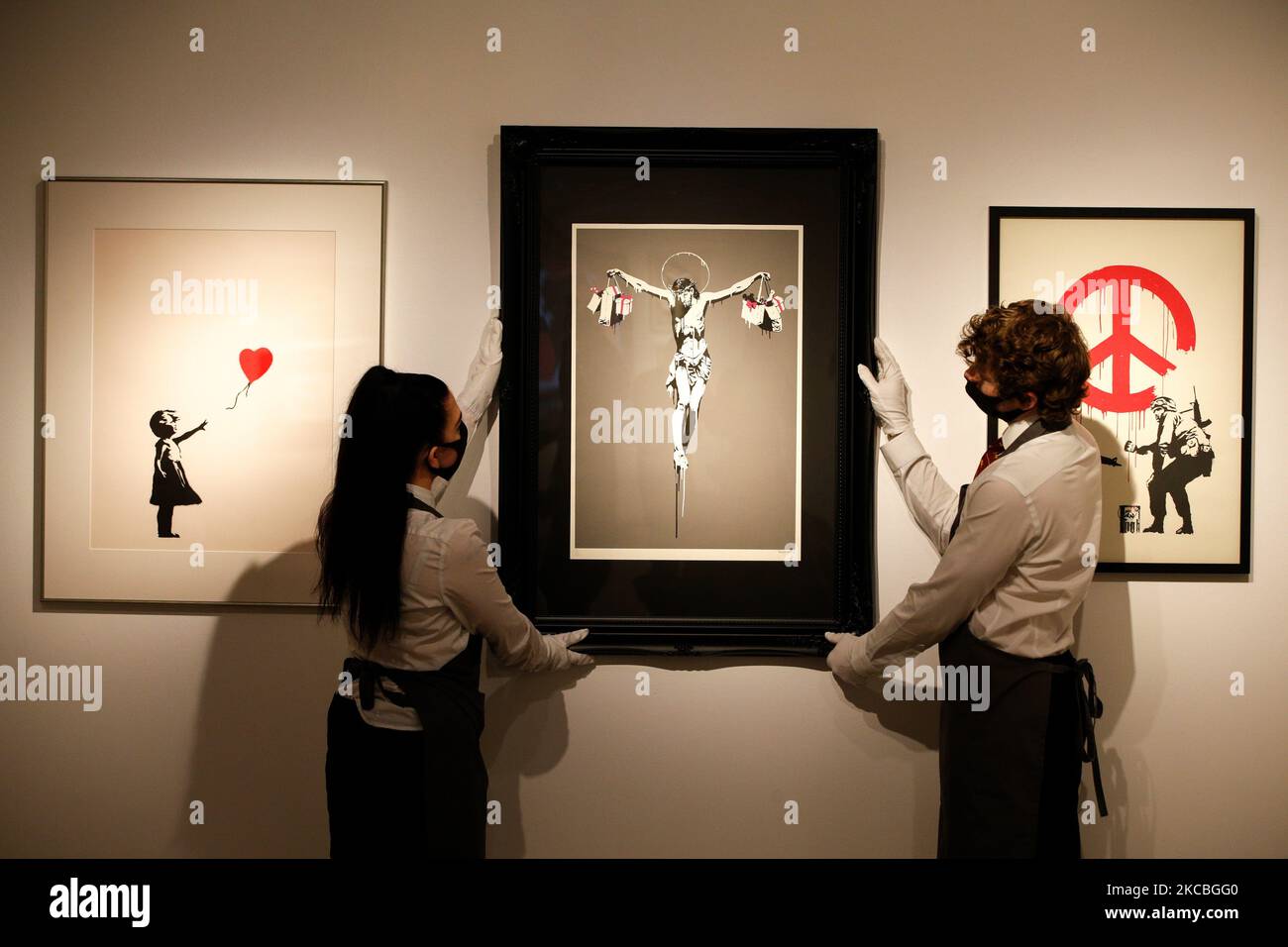 I gestori d'arte posano tenere 'Christ with Shopping', dall'artista britannico Banksy, stimato a GBP100.000-150.000, e tra le opere di Banksy 'Girl with Balloon' (L), stimato a GBP150.000-250.000, e 'CND' (R), stimato a GBP20.000-30.000, durante un'anteprima alla casa d'aste di Christie a Londra, Inghilterra, il 26 marzo 2021. (Foto di David Cliff/NurPhoto) Foto Stock