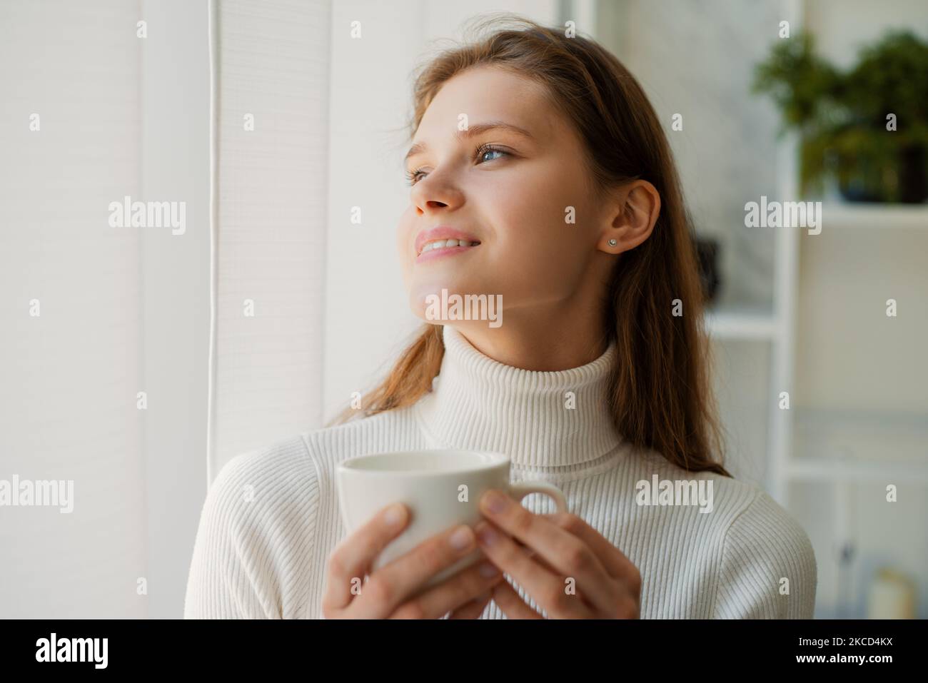 Una giovane donna sorridente che beve il caffè della mattina alla finestra dell'appartamento Foto Stock