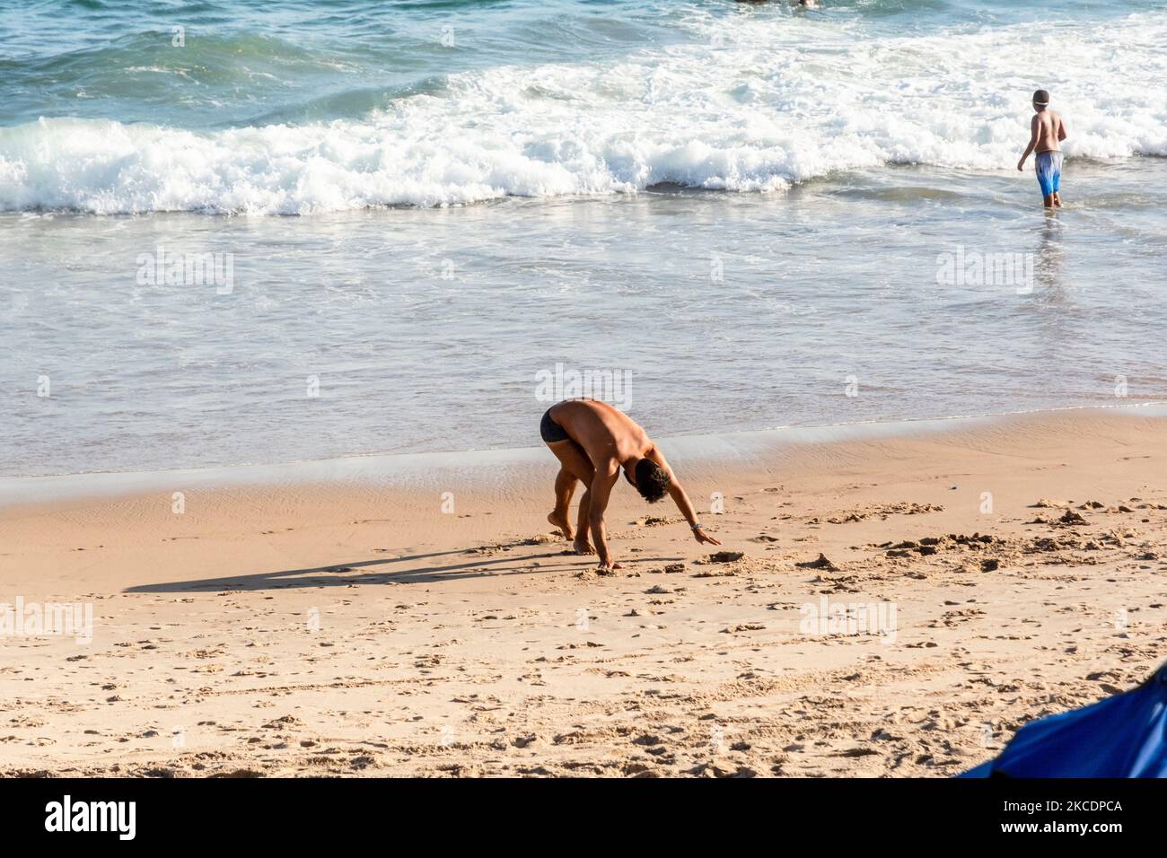 Un uomo fa esercizi di yoga sulla sabbia di Farol da barra spiaggia a Salvador, Brasile, mentre altre persone a piedi Foto Stock