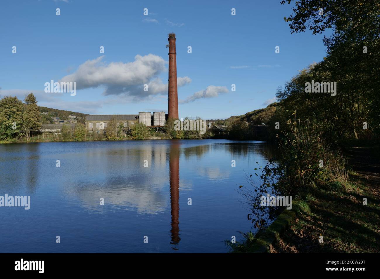 Alto camino di fabbrica di mattoni rossi con riflesso in piccolo lago diga mulino con alberi edifici cielo blu e nuvole bianche Foto Stock