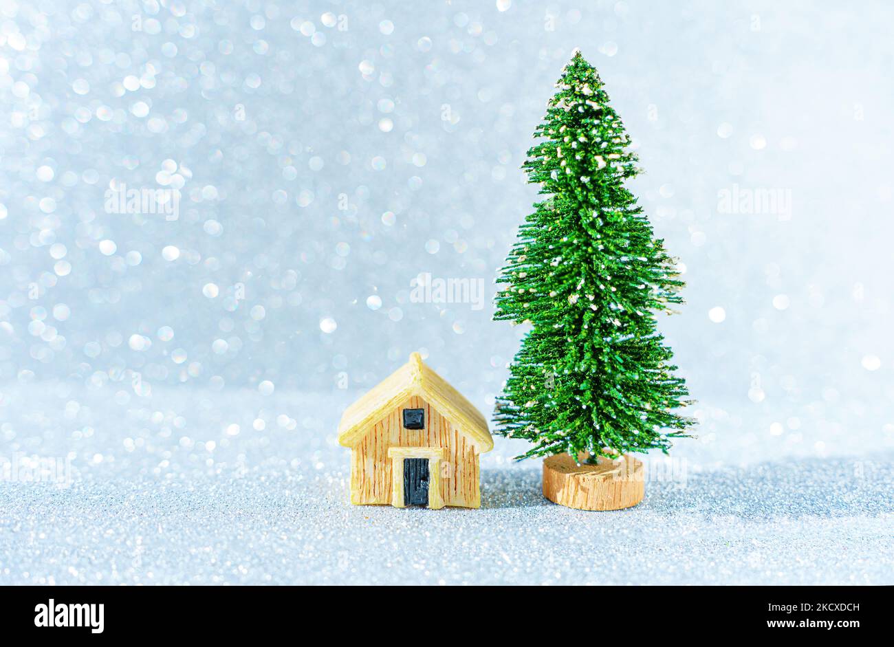 Modello di casa in miniatura posto da un albero di Natale giocattolo su uno sfondo scintillante. Foto Stock