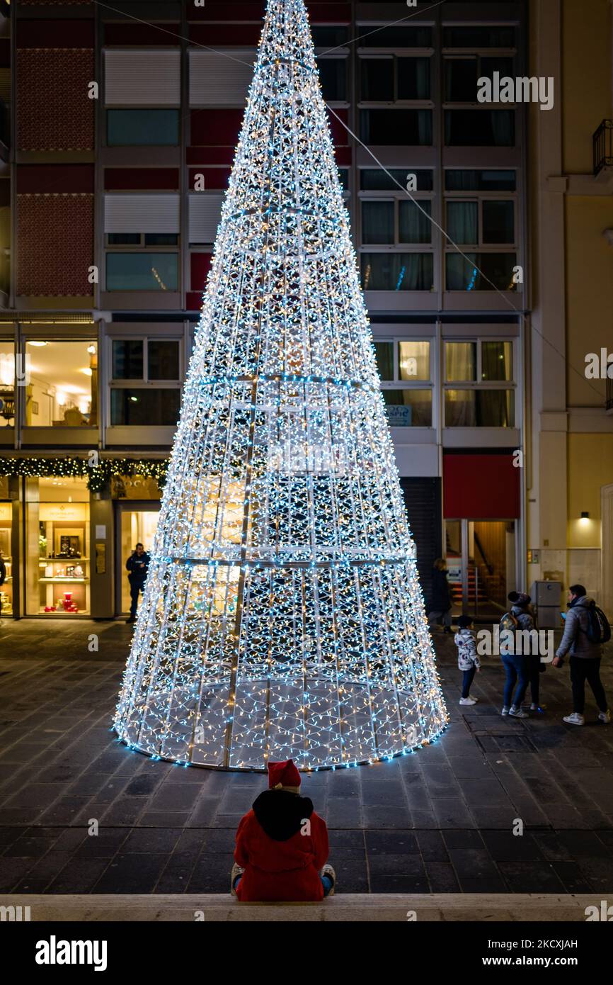 Una bambina vestita come Babbo Natale seduto chiede elemosina di fronte all'albero di Natale nella principale via dello shopping Via Sparano illuminata per Natale a Bari il 9 dicembre 2021. Nonostante la pandemia, il comune di Bari, nel rispetto delle norme di Covid, ha proposto un ricco calendario di eventi per celebrare l'arrivo del Natale e sostenere le attività commerciali in tutta la città. La tradizione inizia il 6 dicembre con l'illuminazione dell'albero di Natale in Piazza del Ferrarese. A pochi passi di distanza il solito legno di Natale istituito da Amgas. Le principali vie dello shopping del c Foto Stock