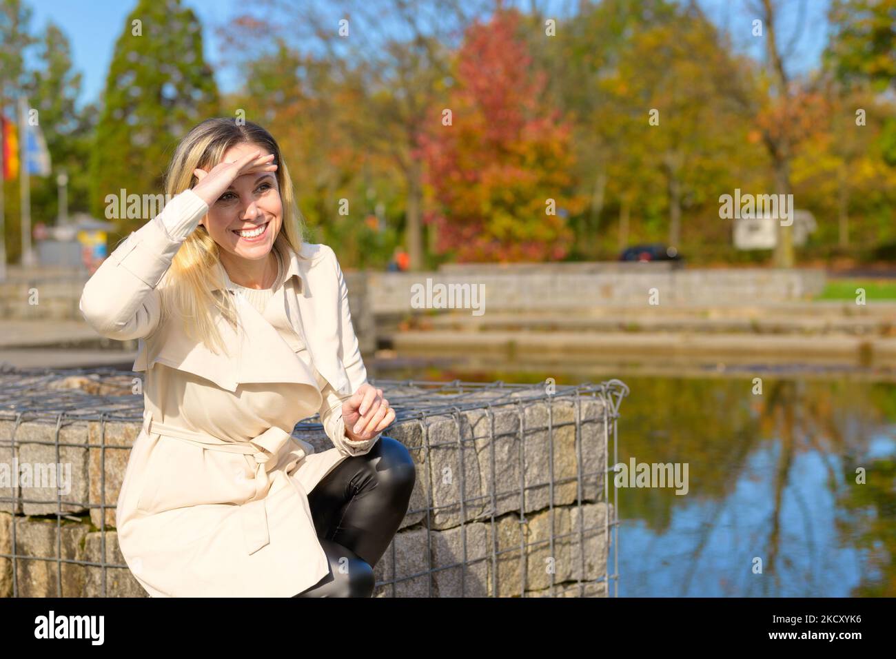 Bella donna bionda di mezza età che si inginocchia vicino a un lago e tiene la mano sopra gli occhi guardando in lontananza di fronte a un parco autunnale Foto Stock