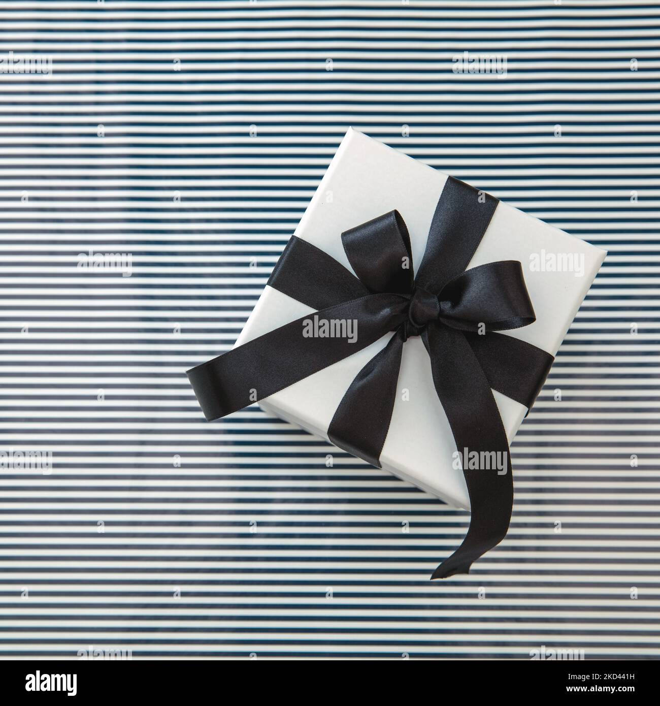 Compleanno anniversario Natale regalo di San Valentino. Una confezione regalo bianca con lussuoso nastro in raso nero su sfondo a righe, vista dall'alto. Foto Stock