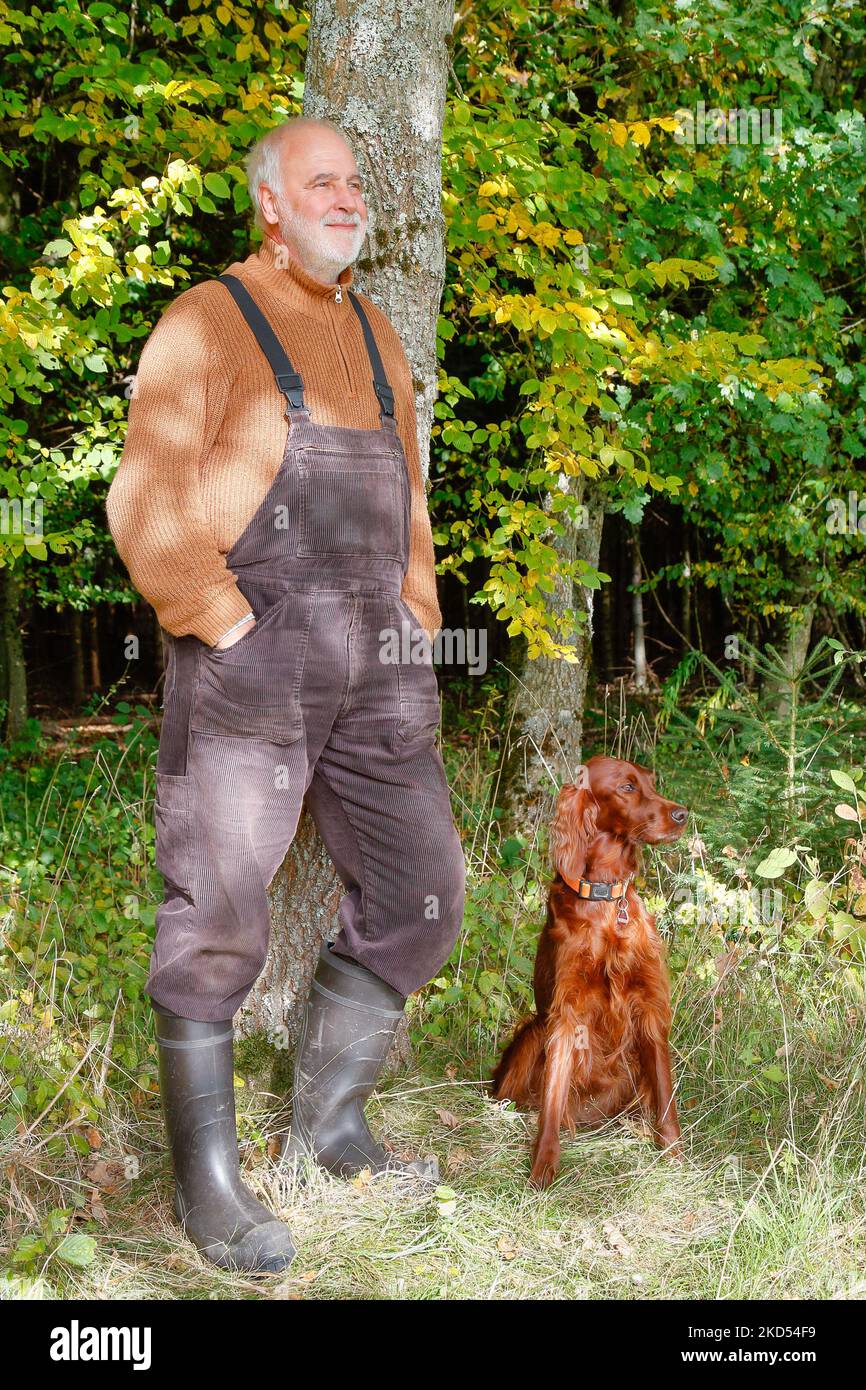 Casualmente in stile country con salopette, maglione, stivali di gomma, un uomo anziano si appoggia contro un tronco d'albero. Foto Stock