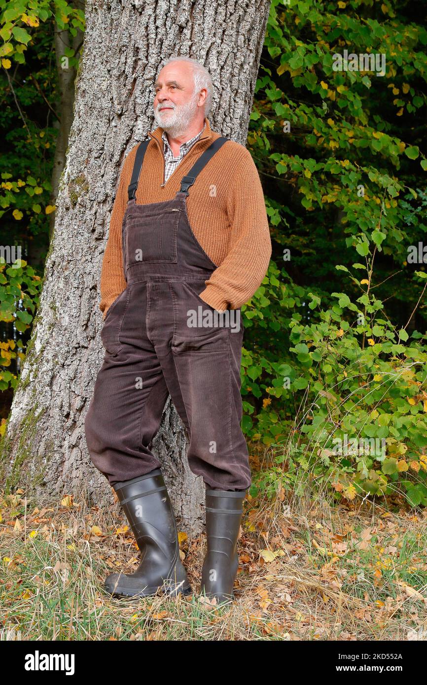 Sorridendo uomo anziano in uno stile casual paese con salopette marrone, maglione e stivali di gomma si appoggia su un tronco d'albero al bordo della foresta. Foto Stock