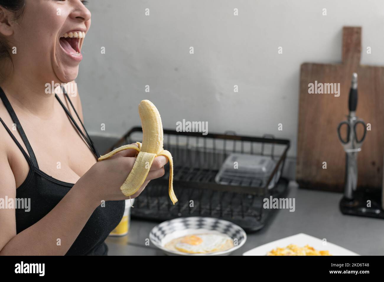bella donna latina con una banana in mano ridendo forte, ragazza mangiare una banana in cucina Foto Stock