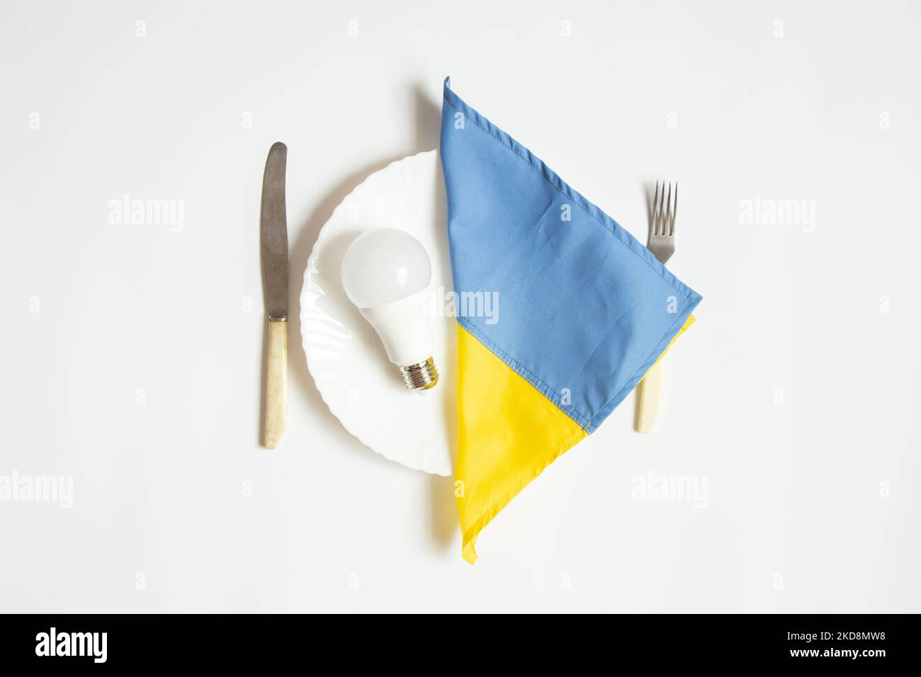 La lampadina a LED e la bandiera dell'Ucraina giacciono su un piatto da pranzo e accanto a un coltello con una forchetta su uno sfondo bianco, in Ucraina si sono avute interruzioni di corrente a causa della Th Foto Stock