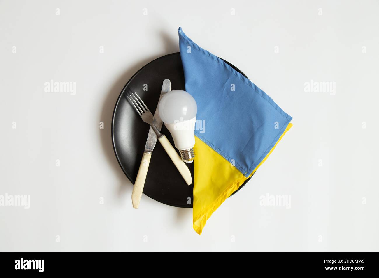 La lampadina a LED e la bandiera dell'Ucraina giacciono su un piatto da pranzo e accanto a un coltello con una forchetta su uno sfondo bianco, in Ucraina si sono avute interruzioni di corrente a causa della Th Foto Stock