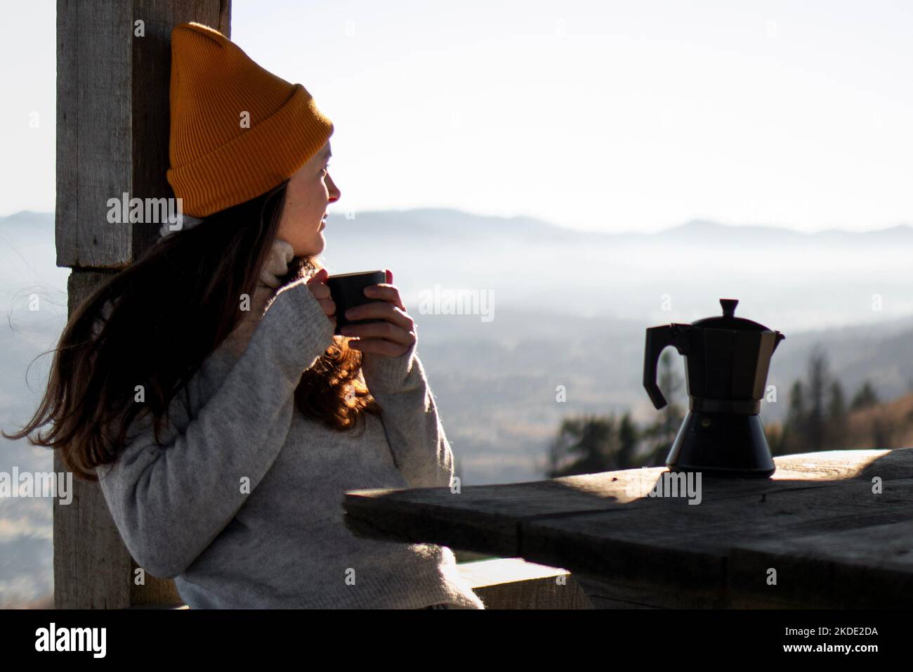 Donna sedersi con una tazza di caffè di fronte a un paesaggio montano e guardare lontano Foto Stock