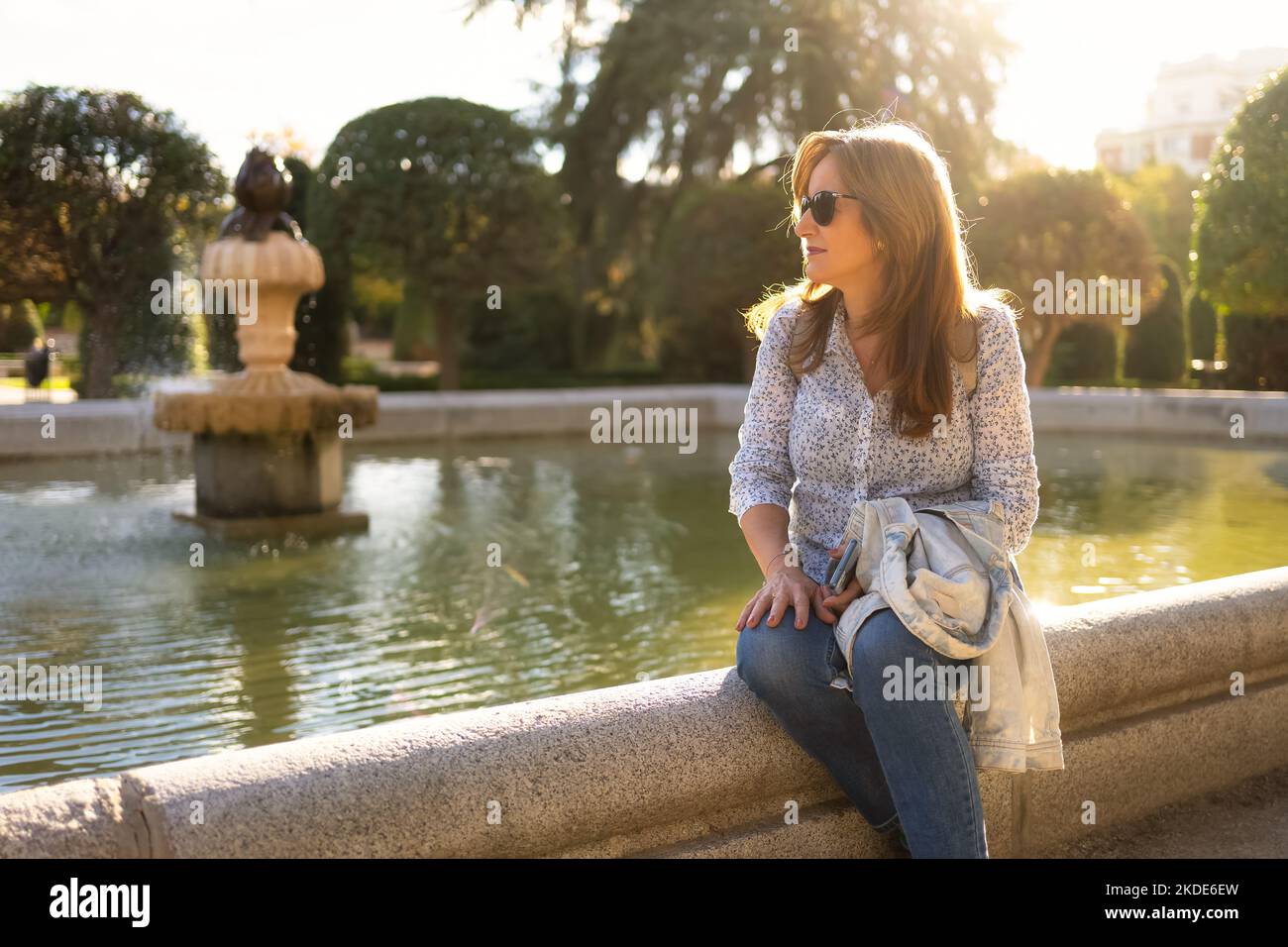 Donna seduta sul bordo di una fontana in un parco pubblico con i raggi del sole che illuminano la scena da dietro. Foto Stock