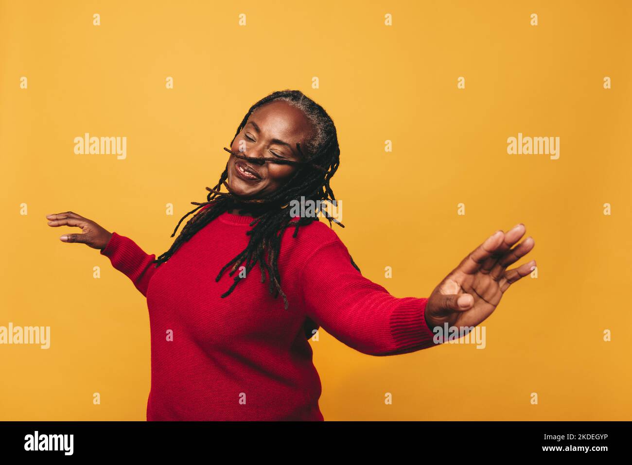 Donna matura gioiosa che balla e si diverte in piedi su uno sfondo giallo. Felice donna nera con Dreadlocks abbracciare i suoi capelli naturali wi Foto Stock