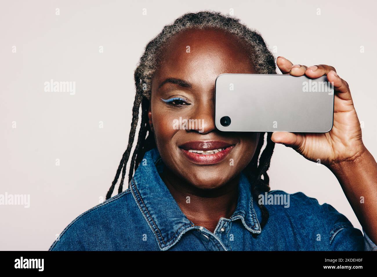 Donna matura che guarda la fotocamera mentre tiene uno smartphone davanti agli occhi. Donna alla moda con lucchetti che indossa una giacca in denim e make-u Foto Stock