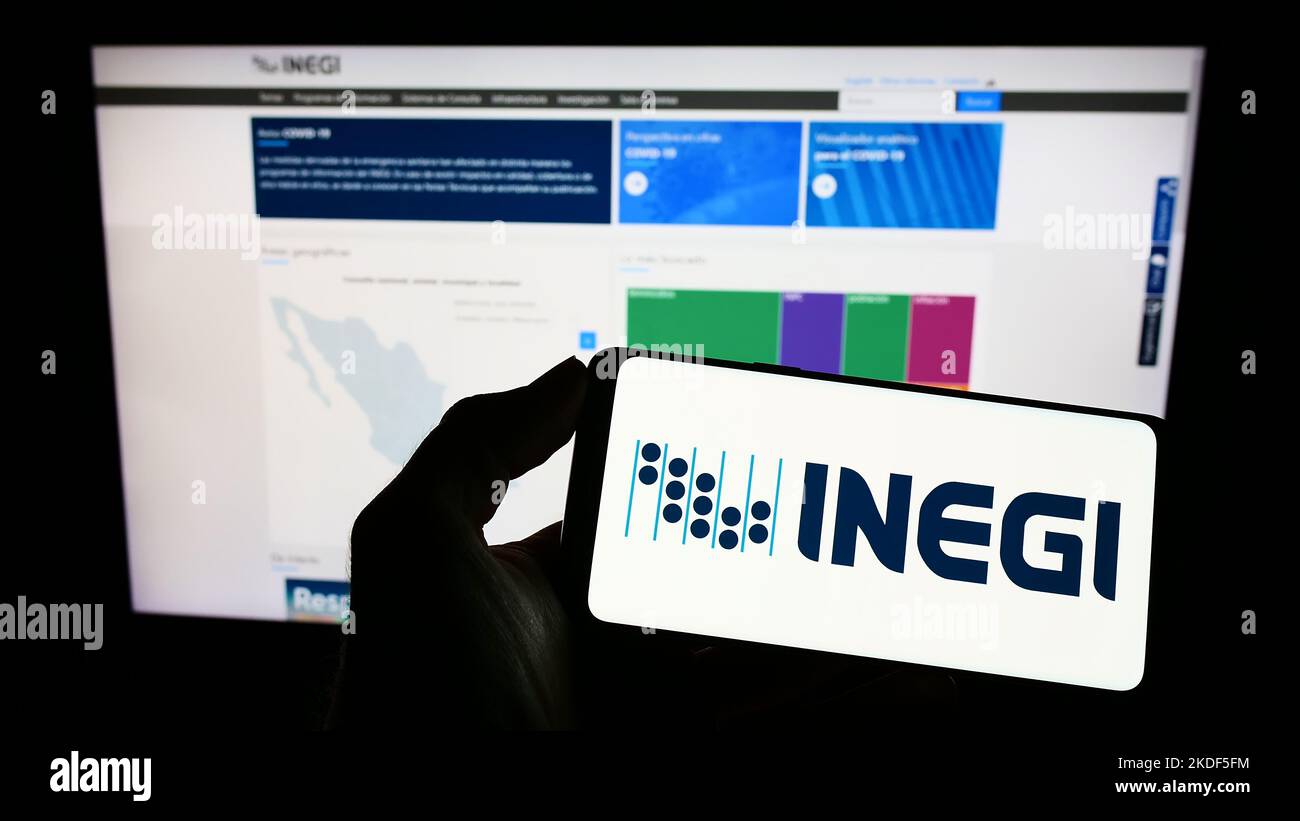 Persona che tiene il telefono cellulare con il logo dell'agenzia di statistiche messicane INEGI sullo schermo di fronte alla pagina web. Messa a fuoco sul display del telefono. Foto Stock