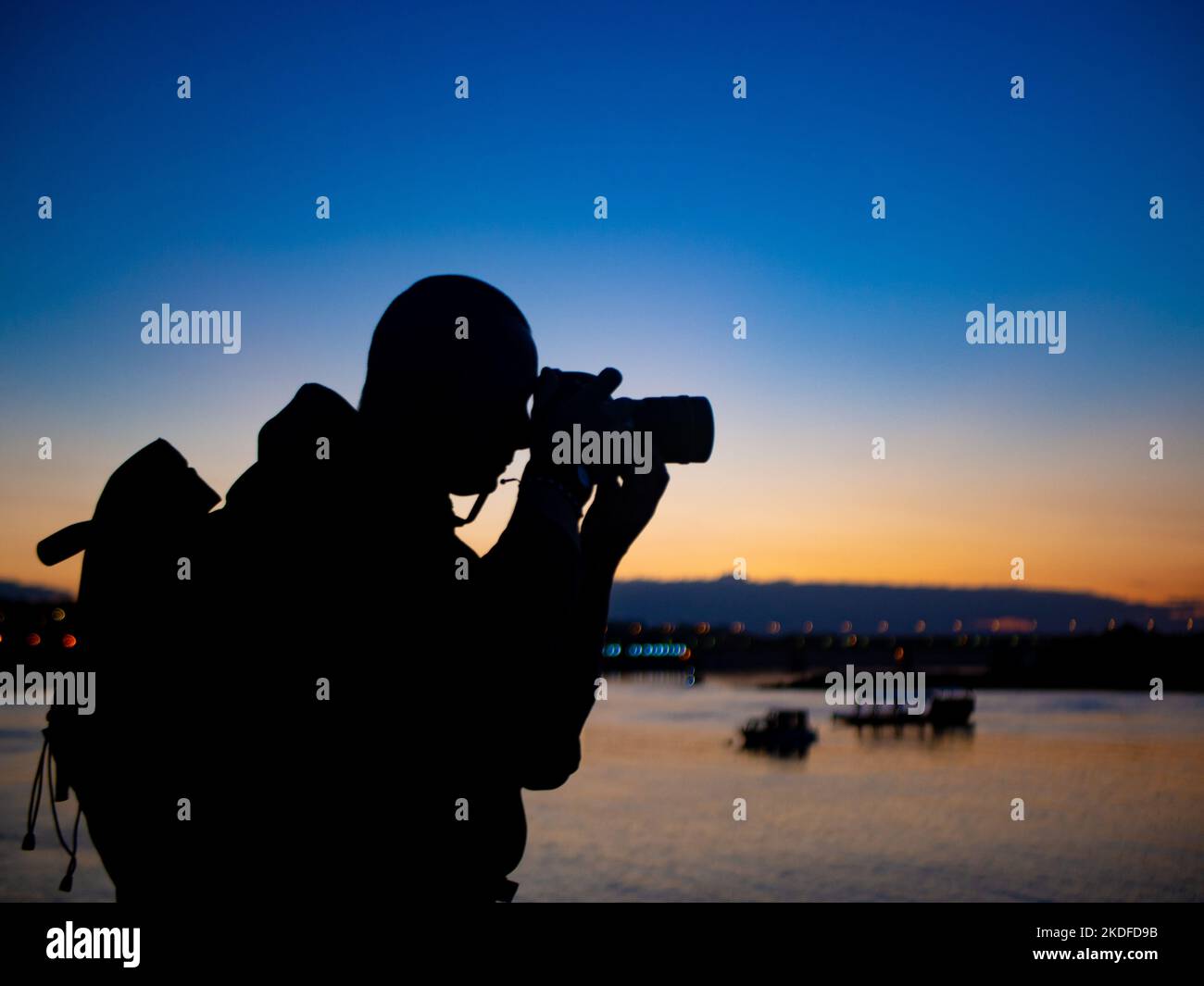 Il fotografo sulla riva del fiume tiene la macchina fotografica al suo occhio e scatta una foto. Crepuscolo, colore di sfondo giallo-blu. Foto Stock