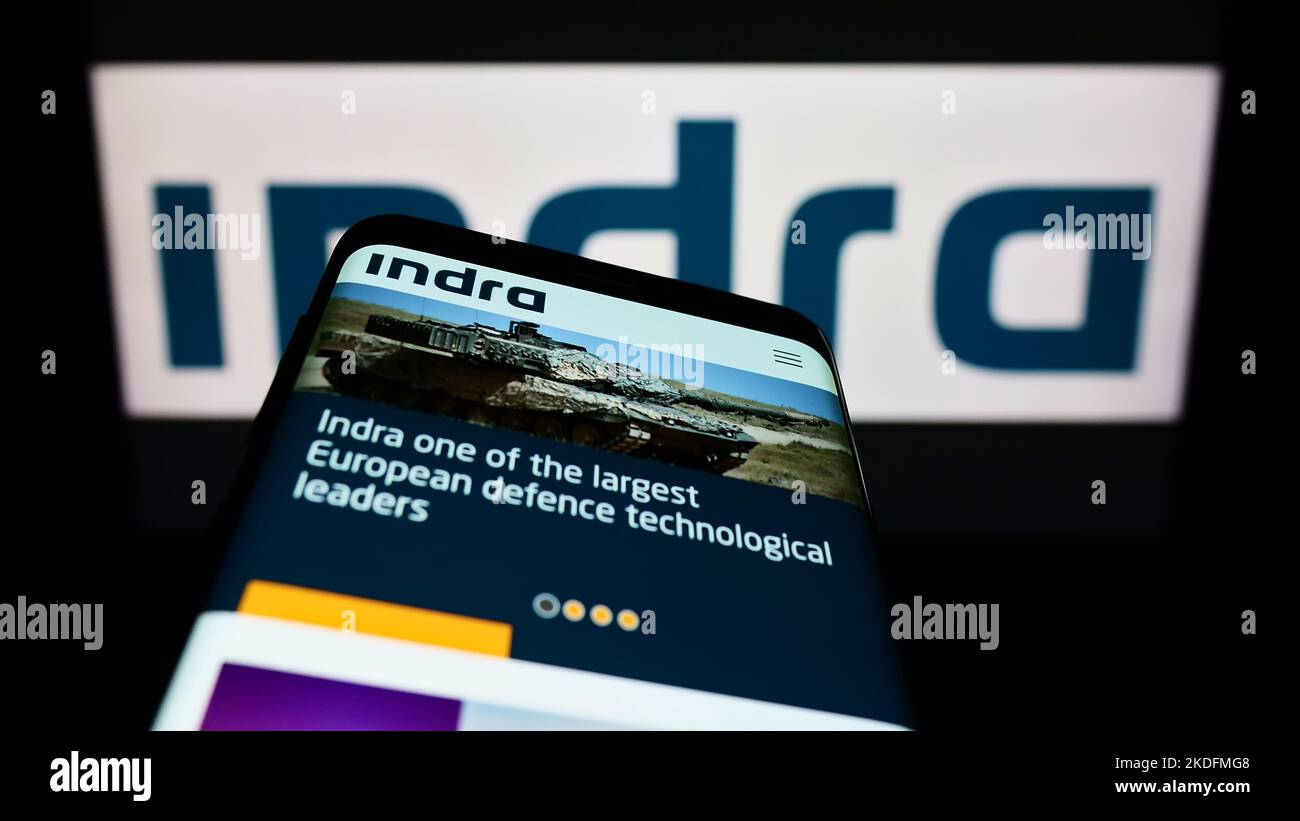 Telefono cellulare con sito web della società spagnola Indra Sistemas S.A. sullo schermo di fronte al logo aziendale. Messa a fuoco in alto a sinistra del display del telefono. Foto Stock