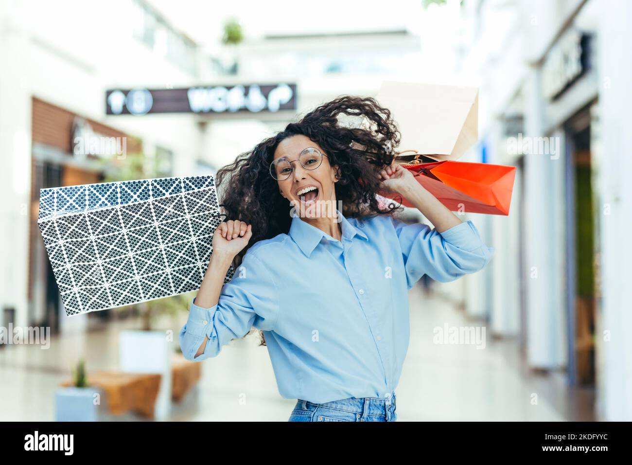 Donna felice acquirente guardando la macchina fotografica e sorridente, donna ispanica con capelli ricci danzando e saltando con piacere, ha acquistato regali in vendita, ritratto di felice donna shopping. Foto Stock