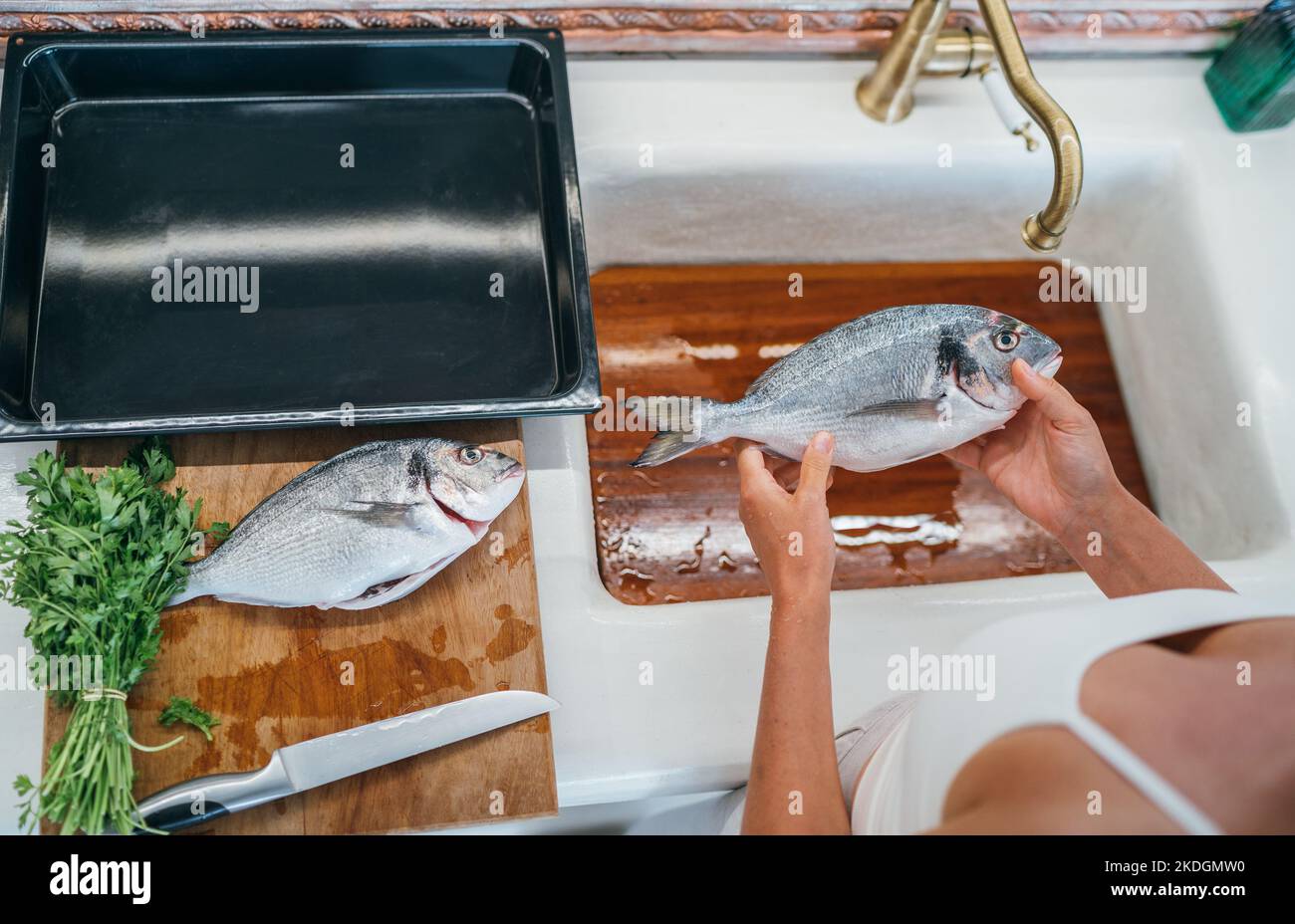Orata fresca o dorada pulizia del pesce dopo la sgrondatura sotto il rubinetto acqua lavandino cucina. Foglio da forno preparato per la cottura. Pesce fresco sano preda Foto Stock